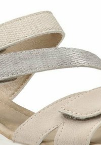 Sandalia beige con tiras cruzadas en textil texturizado y acentos metálicos. Suela blanda y plana con forma curva y cierre ajustable.