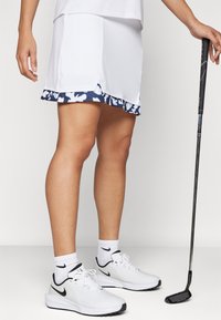 White golf skirt with a navy floral hem, made of lightweight fabric. Paired with white sneakers and a golf club in hand.
