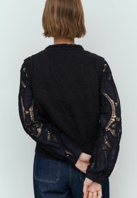 Zwart gebreid sweater met textuur op het lichaam en doorzichtige kantmouwen met ingewikkelde patronen. Donkerdenim jeans zijn gedeeltelijk zichtbaar aan de onderkant.