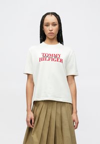 Λευκό βαμβακερό T-shirt με κόκκινο τυπωμένο λογότυπο "TOMMY HILFIGER" στο στήθος. Το μοντέλο φοράει πλισέ χακί φούστα. Ομαλή υφή.