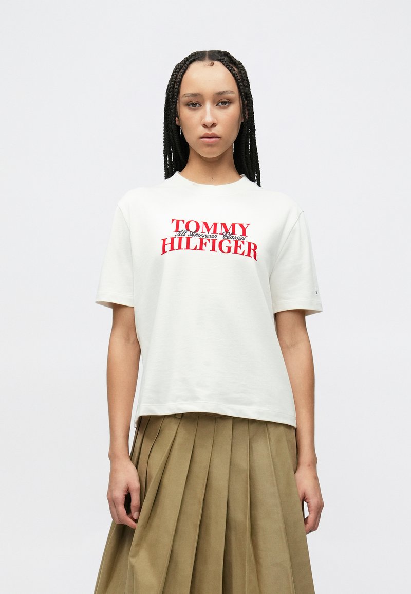 Λευκό βαμβακερό T-shirt με κόκκινο τυπωμένο λογότυπο "TOMMY HILFIGER" στο στήθος. Το μοντέλο φοράει πλισέ χακί φούστα. Ομαλή υφή.