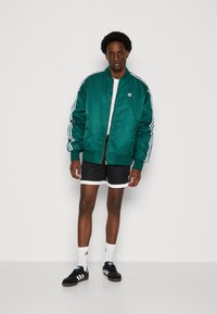 Groene satijnen bomberjack met witte accenten, zwarte shorts met witte details, zwarte sneakers en witte sokken. Model staat tegen een effen achtergrond.