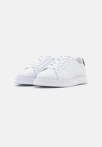 Un par de zapatillas de piel blancas con cordones, suelas gruesas y acentos en verde oscuro en el talón, sobre un fondo blanco.