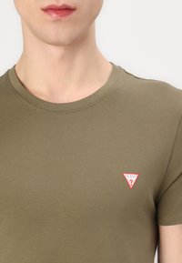 Oliven grøn bomulds t-shirt med rund hals, korte ærmer og et lille rødt og hvidt trekantet logo på brystet. Glat tekstur.