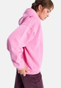 Haut à capuche rose en tissu doux, avec une coupe décontractée, des manches longues, des poignets côtelés et une poche kangourou. Porté avec un pantalon en velours côtelé sombre.