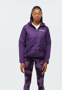 Kvinna klädd i en lila jacka med dragkedja och matchande mönstrade leggings, stående med slutna ögon mot en enfärgad bakgrund.