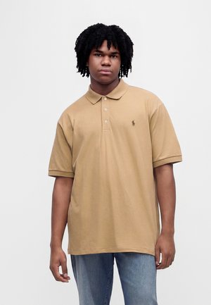 Junger Mann mit lockigem Haar, der ein beiges Polohemd und blaue Jeans trägt, steht vor einem einfarbigen hellen Hintergrund.