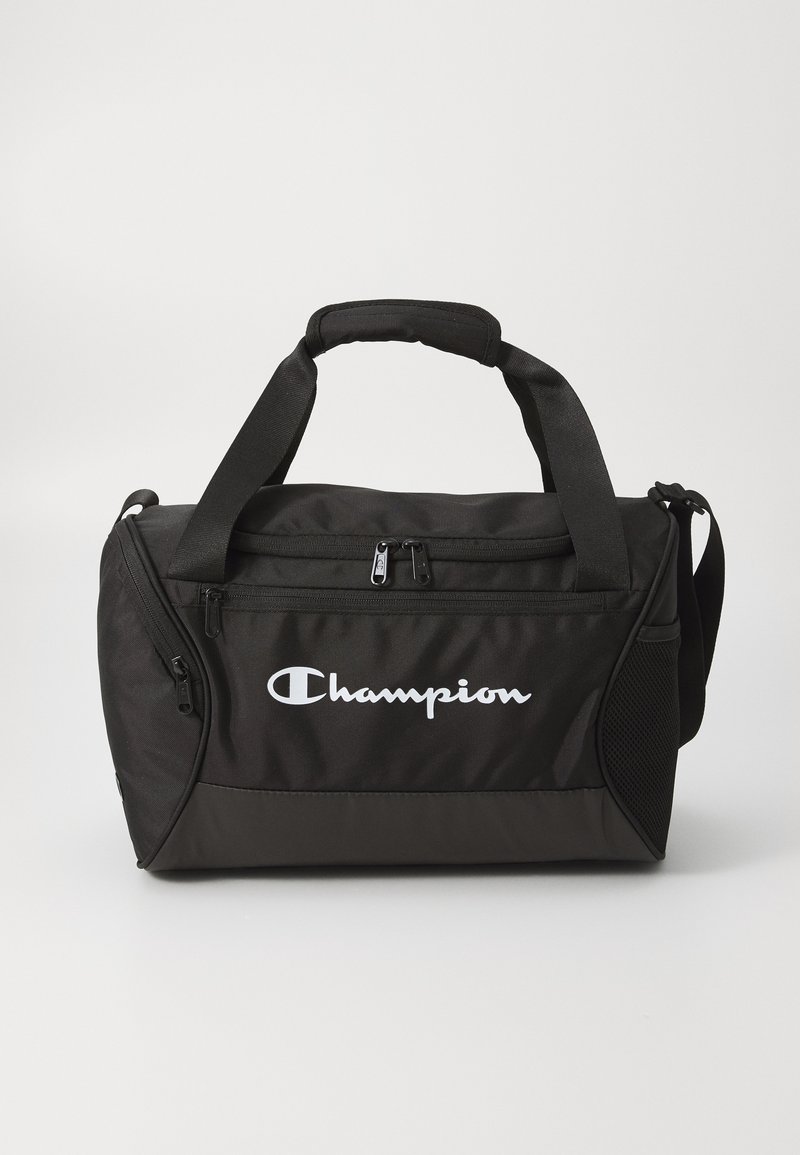 Svart stoff duffelbag med skulderrem, som har en glidelåslukking, meshlomme på siden og en fremtredende hvit "Champion"-logo på forsiden.