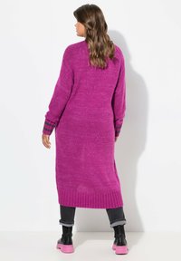 Lange roze sweater met een getextureerde brei, met gestreepte manchetten in donkergroen en zwart. Draagt over grijze jeans en roze schoenen. Achteraanzicht.