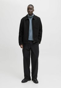 Jonge man die staat, gekleed in een zwart ritsjack, blauwe coltrui, zwarte wijde broek en zwarte chunky schoenen op een witte achtergrond.