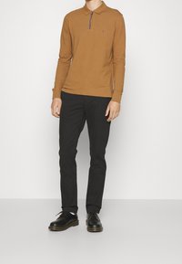 Polo a maniche lunghe marrone con colletto e zip, abbinato a pantaloni neri slim-fit e scarpe nere, su uno sfondo bianco semplice.