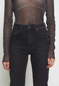 Jean noir taille haute avec une texture lisse, design à cinq poches et fermeture par bouton en métal. Associé à un haut transparent à manches longues scintillant.