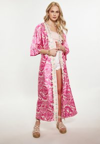 Kimono rosa con motivo a fantasia e maniche ampie, caratterizzato da intricati disegni floreali e una rifinitura a contrasto. Indossato sopra una maglietta leggera e pantaloni corti.