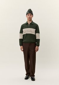 Férfi áll, zöld és bézs hosszú ujjú cipzáras felsőt visel, amin a "Les Deux" felirat látható, barna nadrágot, fekete cipőt és zöld sapkát visel.