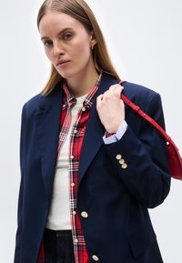 Γυναίκα με ίσια ξανθά μαλλιά φορεί ναυτικό σακάκι πάνω από κόκκινο πουκάμισο με καρό και λευκό τοπ, κρατώντας το λουράκι κόκκινης τσάντας στον ώμο.