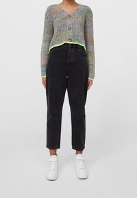 Cardigan lavorato a maglia multicolore con tre bottoni di legno, design corto e bordo verde neon, abbinato a jeans neri a vita alta e sneakers bianche.
