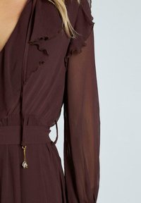 Burgundy klänning med volangkrage, långa genomskinliga ärmar och midjeband med en guldfärgad detalj som har en hängande pärla. Strukturerad tyg synlig.