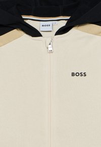 Φούτερ με κουκούλα και φερμουάρ σε κρεμ, μαύρο και μπεζ. Διαθέτει εμπρόσθιο φερμουάρ, ριμπ υφή και λογότυπο "BOSS" σε μαύρο χρώμα στο στήθος.