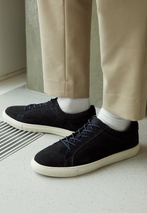 Fekete velúr sportcipők kék fűzőkkel, fehér gumitalppal és texturált felülettel, bézs nadrággal és fehér zoknival viselve, gyöngyözött padlón.