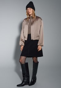 Licht taupe satijnen bomberjack, zwarte getailleerde rok, gebreide zwarte beanie en zwarte leren laarzen tot aan de knie. Soepele texturen en een minimalistisch ontwerp.