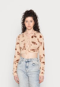 Cardigan cropped em bege tricotado com padrões de folhas castanhas, mangas longas e abertura frontal com cordões. Combinado com jeans claros de cintura alta.
