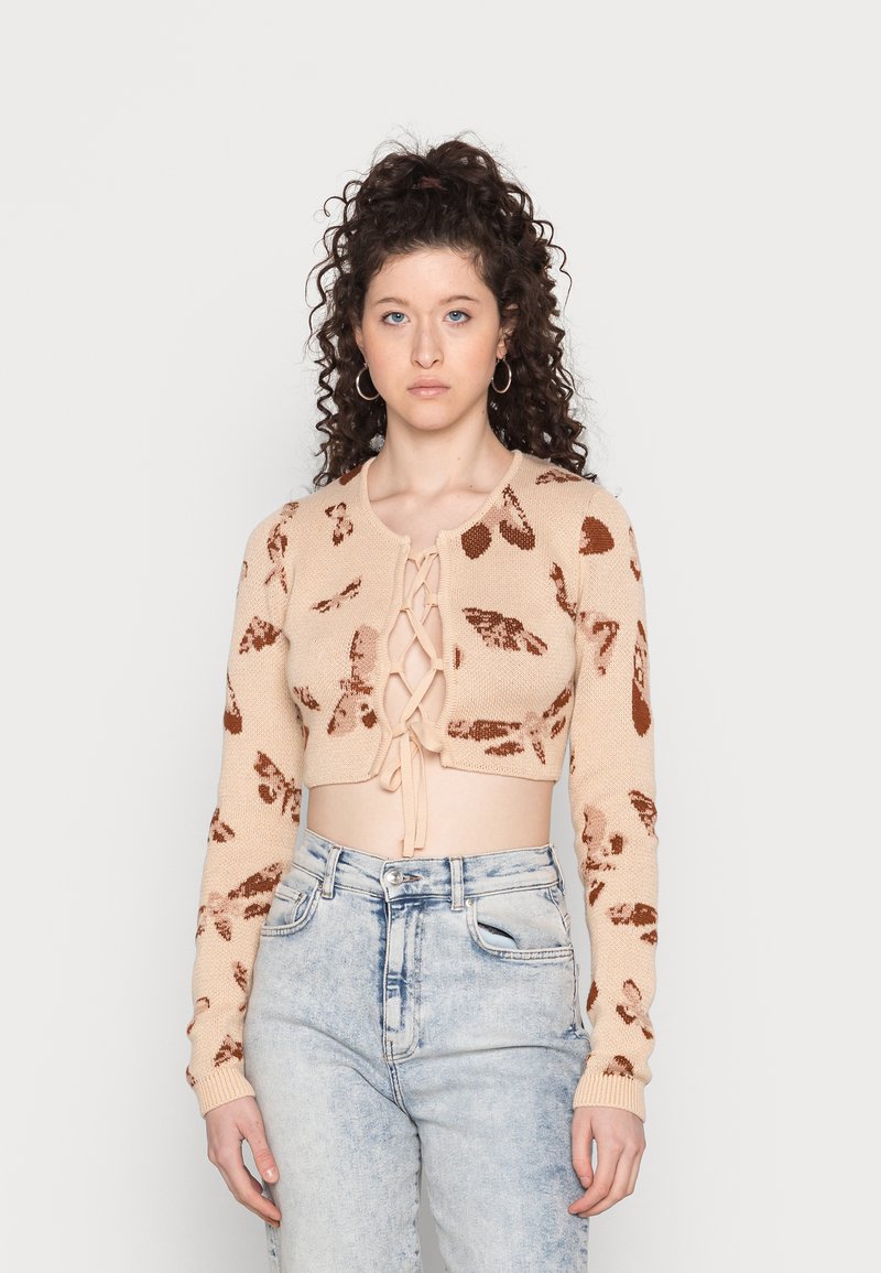 Cardigan cropped em bege tricotado com padrões de folhas castanhas, mangas longas e abertura frontal com cordões. Combinado com jeans claros de cintura alta.