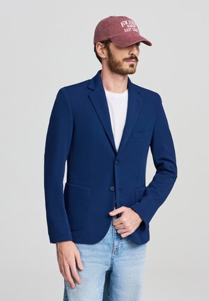 Blazer blu navy con due tasche anteriori, chiusura a un bottone e rever a pattina, abbinato a una t-shirt bianca e jeans azzurri chiari.
