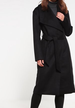 Femme portant un manteau en laine noir ceinturé sur un pull à col roulé gris foncé, debout devant un fond blanc uni.