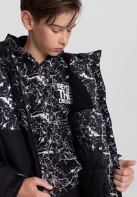 Młody chłopiec w czarno-białym wzorzystym hoodie i dopasowanej kurtce, patrzący w dół, trzymający kurtkę otwartą obiema rękami.