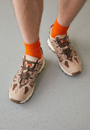 Personne portant des baskets Adidas beige et marron avec des chaussettes chevilles orange vif debout sur un sol gris clair tacheté.