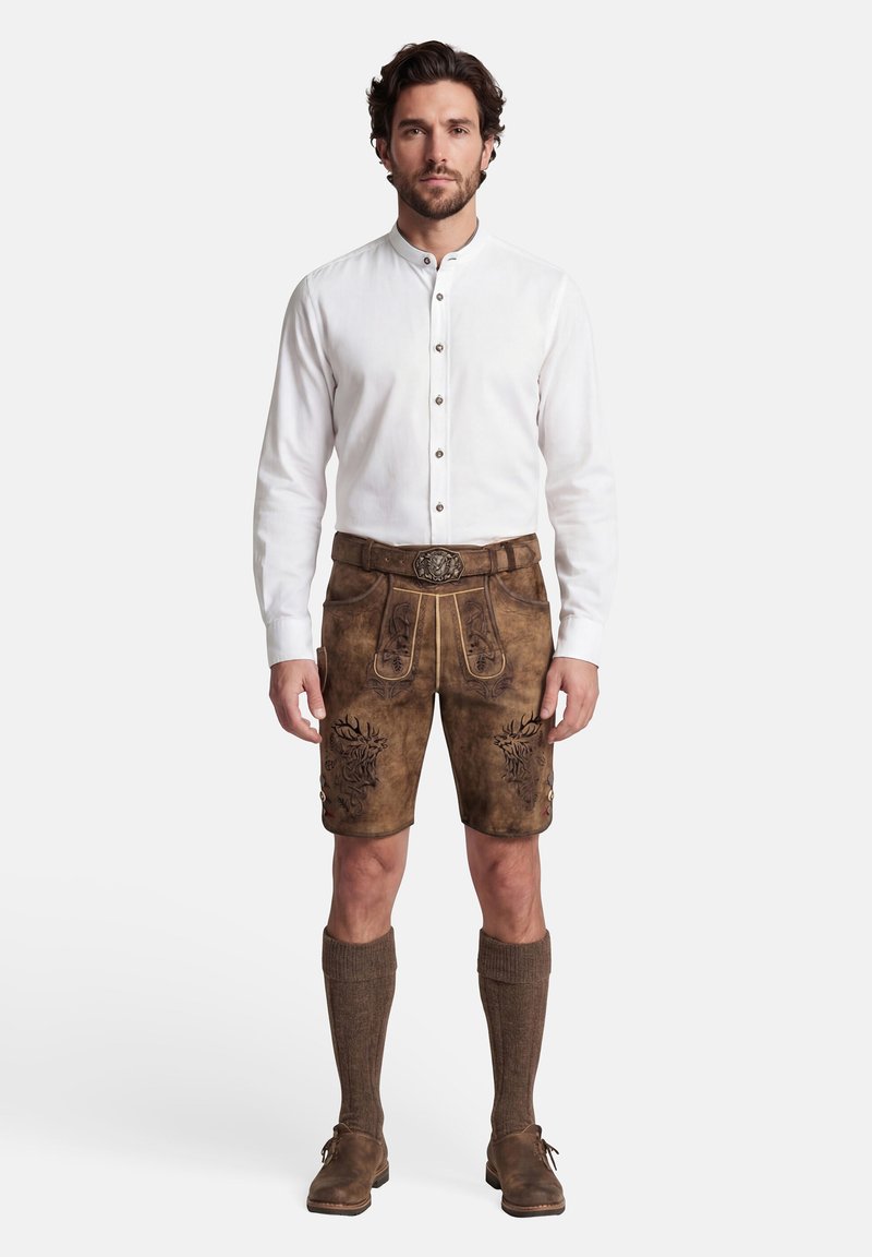 Mann trägt traditionelle braune Lederhose mit bestickten Details, weißes Hemd mit Knöpfen, braune knielange Socken und braune Schuhe, steht.