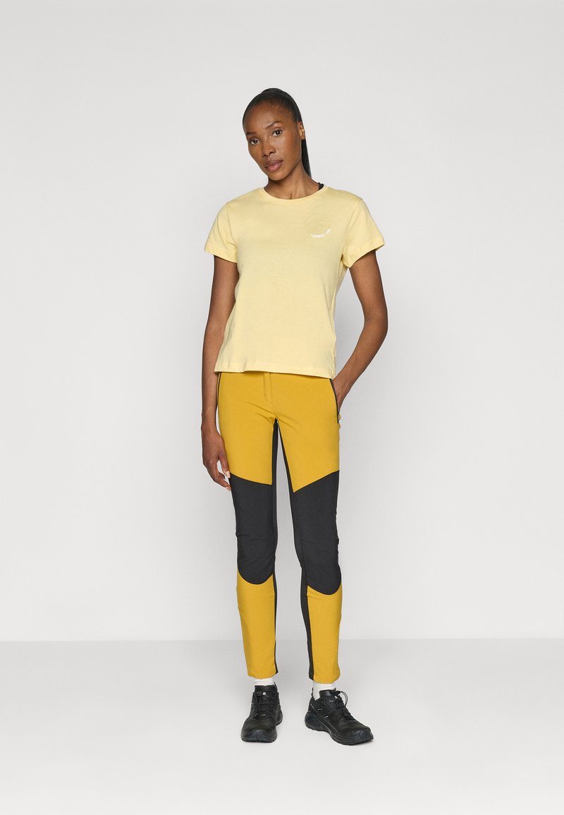 Żółta bawełniana koszulka z okrągłym dekoltem i krótkimi rękawami, w połączeniu z legginsami w żółto-czarną kolorystykę i czarnymi butami sportowymi.