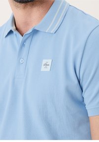 Světle modré polo tričko s texturovanou látkou, má dvoutlačítkovou légu a kontrastní límec s bílými pruhy. Obsahuje značkovou etiketu.