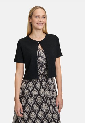 Cardigan noir à manches courtes avec un col rond et une fermeture à un bouton à l'avant, porté par-dessus une robe à motifs.