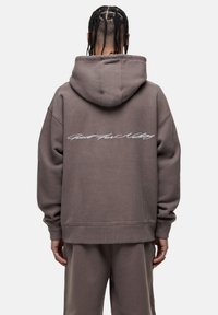 Felpa oversize con cappuccio in marrone tenue con finitura testurizzata. Presenta testo corsivo bianco sulla schiena e polsini a costine.