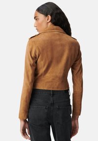 Mujer con cabello largo y oscuro, vestida con una chaqueta de ante color beige recortada y jeans negros de cintura alta, de espaldas contra un fondo blanco.