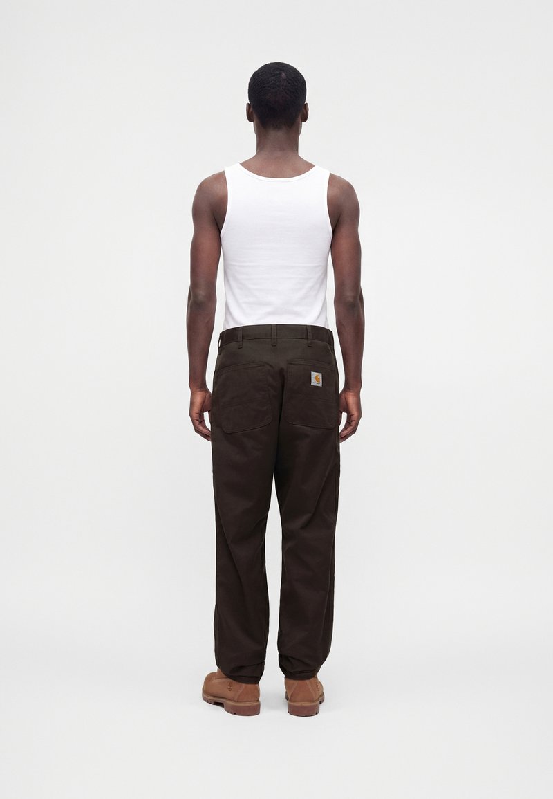 Homme portant un débardeur blanc, un pantalon marron foncé avec un patch logo sur la poche arrière, et des bottes de travail beige, debout de dos.