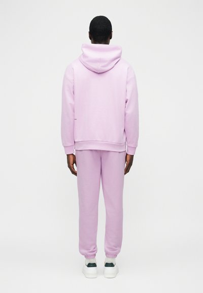 Háttal látható személy, aki világoslila kapucnis pulóvert és hozzáillő melegítőnadrágot visel fehér sportcipővel, egyszerű fehér háttér előtt.