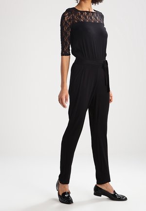 Femme portant une combinaison noire avec des manches en dentelle trois-quarts, une taille ceinturée et des mocassins en cuir verni noir, debout devant un fond uni.