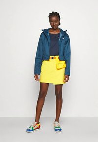 Kék vízálló dzseki, tengerészkék felső, sárga szoknya oldalzsebbel, színes tornacipők. Rövid fazon, testhez simuló sziluett és cipzár.