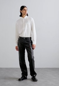 Biely button-up košeľový s kapsou, spárovaný s čiernymi textúrovanými koženými nohavicami. Model má na sebe čierne topánky a stojí pred jednoduchým pozadím.