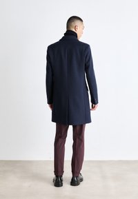 Marineblå ullfrakk med høy krage og slank passform, med en rett søm på baksiden, kombinert med burgunder bukser og svarte sko.