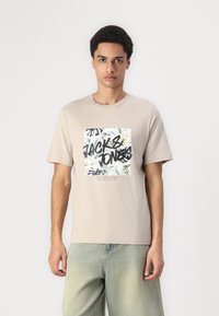 Camiseta beige de manga corta con un gráfico cuadrado que presenta hojas tropicales y texto en negro, combinada con pantalones cortos de mezclilla clara.