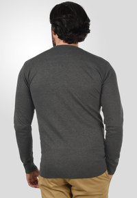 Pull en tricot gris à manches longues, col rond et poignets côtelés. Texture lisse. Porté sur un pantalon de couleur claire, vu de derrière.