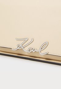 Borsa in pelle beige con logo "Karl" argentato e lucido. Presenta dettagli neri e cuciture visibili, evidenziando una texture liscia e una forma strutturata.