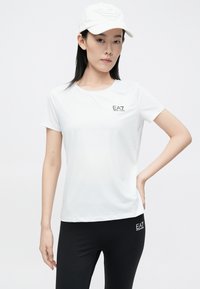 Fehér sportos rövid ujjú póló kerek nyakkal, bal mellkason nyomtatott EA7 logóval, fekete leggingsszel párosítva, amely logó részlettel rendelkezik.