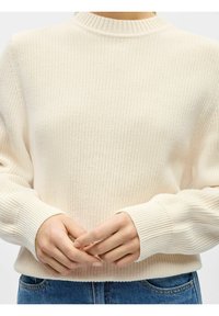 Neulepusero, joka on kermaan vivahtavan värinen ja jossa on resorit korkea kaulus ja hihansuut. Siinä on tekstuuri kuvio ja rento istuvuus. Kädet on asetettu vyötärölle.