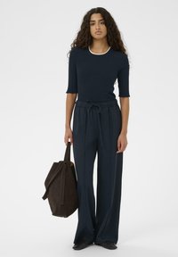 Vrouw met lang krullend haar draagt een marineblauwe geribbelde top en wijde broek, en houdt een grote donkerbruine tote bag tegen een witte achtergrond.