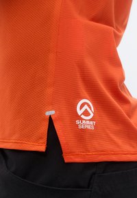 Κοντινό πλάνο στο τελείωμα πορτοκαλί αθλητικής μπλούζας με υφή και λογότυπο «Summit Series», φορεμένη πάνω από μαύρο παντελόνι.