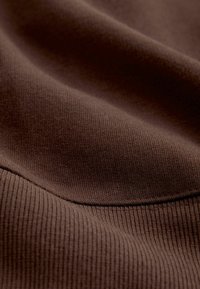 Hnedá ribbovaná tkanina s jemnou textúrou, s miernym natiahnutím a jemnými záhybmi. Povrch vykazuje konštantnú farbu a vzor.